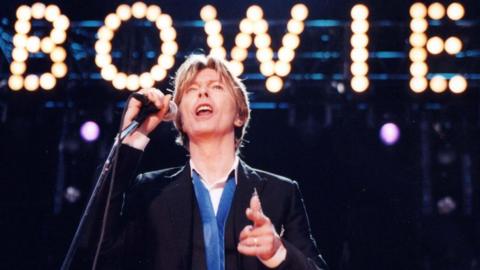 David Bowie, wearing a suit jacket and blue and white shirt, performs on stage with a sign saying BOWIE behind him