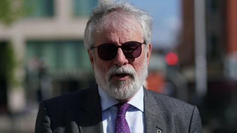 Gerry Adams with grey hair and sungalsses. He is wearing a grey suit, blue shirt and purple tie. He is outside.
