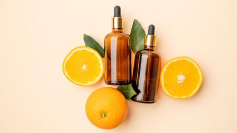 Two slices of oranges and one whole orange and placed around two brown bottles with pipettes in them. Suggestion is that the bottles contain vitamin c face serum which is frequently used to help skin improve. 