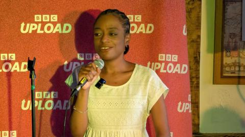 A woman talks into a microphone on stage. She stands in front of a red backdrop which says "BBC Upload" on it.