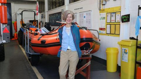 A woman with bobed hair and a pale blue cardigan stands in front of a bright orange lifeboat