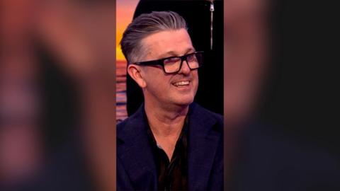 David McCormack, the voice of Bandit Heeler, smiles. He wears black glasses and has swept back grey hair.