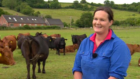 A farmer - a woman in her 40s wearing a blue shirt with rolled up sleeves - stands in a field surrounded by calves on a gloomy day. A farmhouse can be seen in the distance.