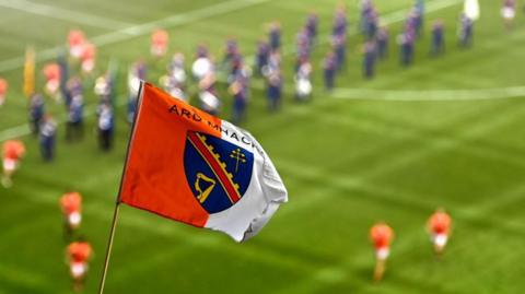 An Armagh GAA flag flies in the foreground - in the blurred background players in orange Armagh jerseys can be seen on a grass pitch, as well as members of a marching band. The flag is orange and white with a blue crest in the middle.