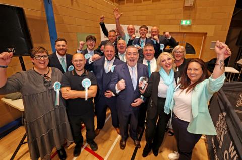 Reform councillors celebrate in Wigan. They wear pale blue rosettes and wave their hands in the air 