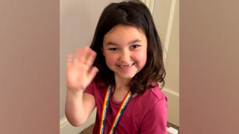 Bebe is pictured sitting in a little white wooden chair. She is smiling and waving with her right hand. She has long brown hair and is wearing a pink top and pink patterned leggings.