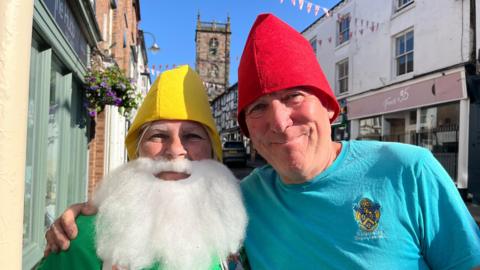 A woman and man with their arms around each other and smiling. The woman, Cllr Sue Sealy, is on the left of the photo - she is wearing a bright yellow pointed hat, a false white beard, and a green top with grey braces. The man, Morris Clive, is wearing a bright blue tshirt and a red pointed hat. They are stood on the high street, with a church spire behind them and bright blue skies.