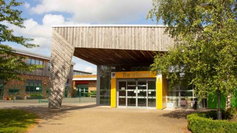 The entrance to a modern school building. Columns are painted yellow around a glass door, with a large wood clad pillar and roof. Willows is written in silver letters above the door, with reception written on the glass. Other school buildings can be seen to the left, with trees and bushes lining the path.