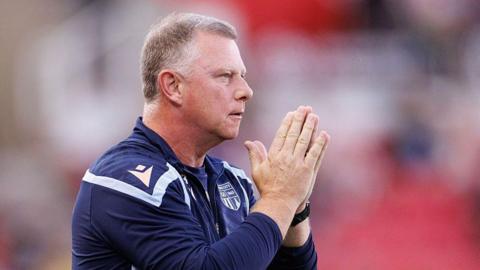 Stoke City boss Mark Robins watches from the touchline. He is wearing a club sweat-top and has his hands together looking a bit like he is saying a prayer.