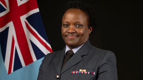 Joan Ochuodho has black hair and is wearing a navy uniform jacket with military stripes, a black tie and a blue shirt. She is standing in front of the Union flag.