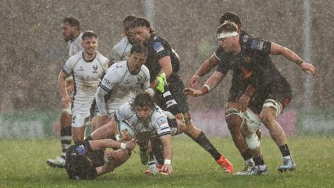 Bernhard Janse van Rensburg powers forward in a rainstorm as various Bristol Bears and Exeter Chiefs players look on.