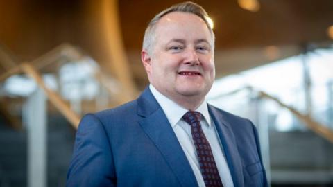 Darren Millar stood inside the Senedd building, wearing a blue suit, tie and white shirt.