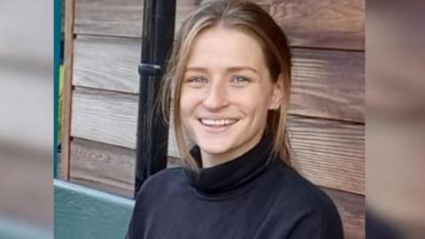 A young woman with long hair in a pony tail and wearing a black turtle neck smiles at the camera. She is sitting in front of a shed.