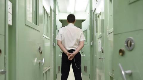 A member of prison staff stands with bag to camera, flanked by closed prison cells