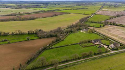 Aerial view of green fields lined by trees and paths. A couple houses can be spotted among the fields.