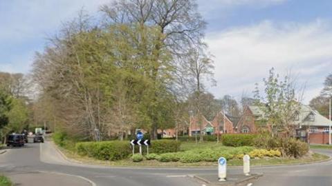A google maps image of a roundabout. There are signs telling drivers to drive to the left. There are bushes and trees on the roundabout. There are some houses in the background.
