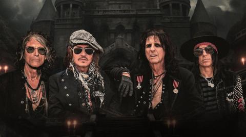 A group of four men dressed in dramatic rock‑and‑roll attire stand in front of a dark, gothic castle with tall spires. They wear sunglasses, hats, scarves, jewellery, and studded jackets, with the central figure in a medal‑covered coat.