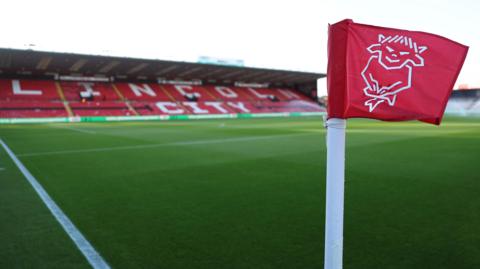 A red corner flag at Lincoln City's LNER Stadium with the club's Imp logo in white. The pitch and the red seats of the GBM Stand with Lincoln City written in white seats can be seen behind.