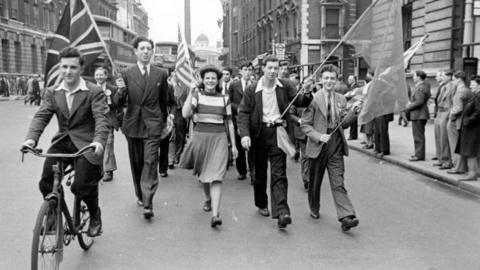 8th May 1945: Students from London University stage an impromptu VE Day parade. Men in suits are marching on a street towards the camera. One man at the front on the right is riding a bicycle. Three men and a woman are waving Union Jack flags.