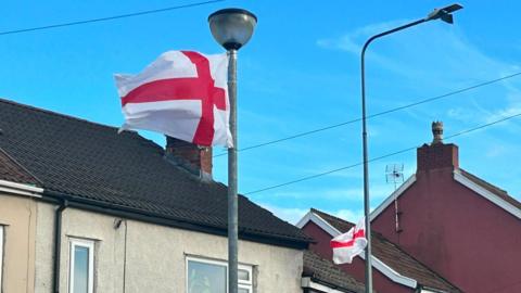 England flags attached to two lampposts down a residential street. Houses can be seen behind them.