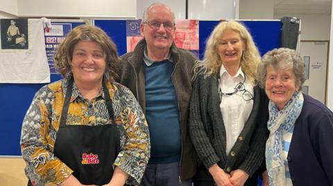 Smiling Carole Anne Robins is wearing an apron. She is with her husband Roger Rondel Donna Murphy, the senior chargehand porter at Jersey General Hospital and Eileen Palmer who are standing in a hallway at the hospital.