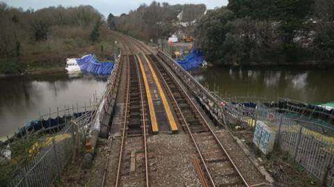 A railway bridge over a river. There are trees and some buildings on either side of the tracks beyond the bridge.