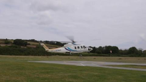 A helicopter hovering above a tarmac square in a green field, with trees behind. 