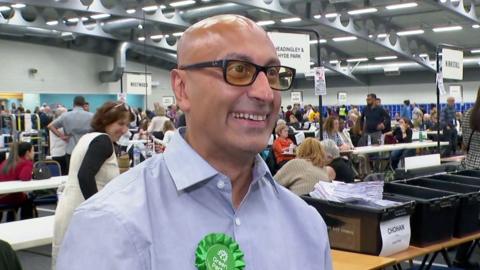 A bald man wearing tinted yellow glasses and a blue shirt wears a green rosette. He is smiling 
