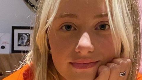 A girl called Kezia Mason. She has long, straight blonde hair. Her face is leaning against her left hand, which has a silver ring on her middle finger. She has a small smile on her face.