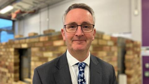 Close-up image of Simon Cook, principal of Mid Kent College. He is smiling and wearing a grey jacket, white shirt, blue tie and glasses. He is standing in a room in front of a wall of bricks.