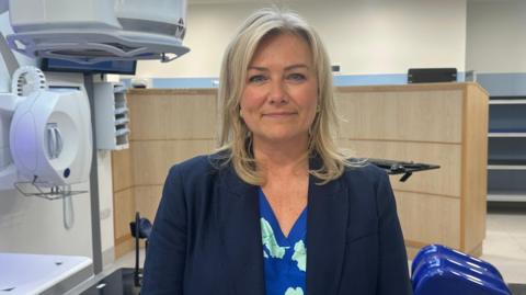 A smiling woman stood in front of a white piece of hospital equipment. She has shoulder-length, blonde hair and is wearing a blue suit jacket with a blue top underneath which has a floral pattern on. 
