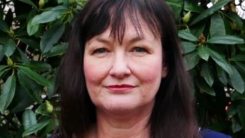 Head and shoulders image of Lynn Kelly, who has long dark hair and is standing in front of a rhododendron