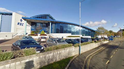 The exterior of the National Sports Centre, which is a large blue and white glass-fronted building with a slopping roof. There are cars parked in front of it and a white wall partitions the car park from a bus stop on the road alongside the complex.