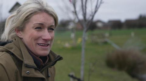 Farm manager Kirsty Cropper with blond hair and green wax waterproof coat smiling with the trees planted in the background 