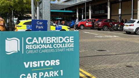 A police car is parked at the college behind a blue sign that reads "Cambridge Regional College - visitors car park 1".