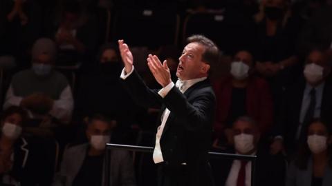 James Ross in formal concert attire stands on a podium with both hands raised during a performance in a concert hall. The conductor is illuminated by a spotlight in the foreground, while rows of audience members sit in the dimly lit background. Many audience members are wearing face masks.