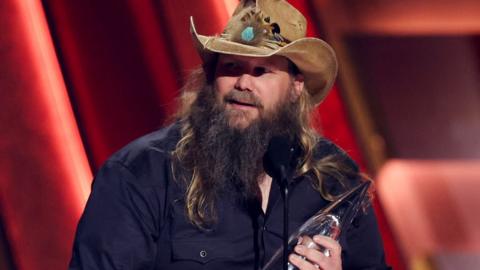 Chris Stapleton with hat, beard and long hair, holding a CMA Award