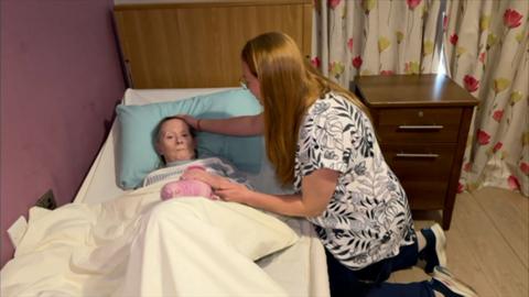 A woman lying in a care home bed with her daughter, a woman with a black and white patterned top and long red hair, by her side