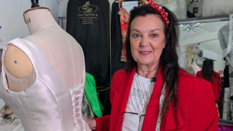 In this picture Angie is smiling at the camera as she stands next to a mannequin. Angie wears a red blazer and a white t-shirt.  She's also wearing a sparkly red headband and has long black hair. 