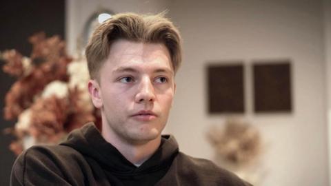 A young man with middle parted, cropped light blonde hair looks across the frame. He is wearing a black hoodie and is sat in front of a vase of flowers in an indoor area.