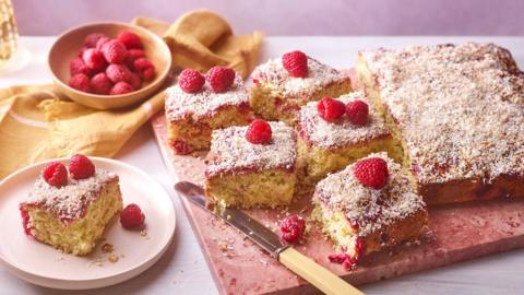 Image of raspberry and coconut cake, cut in squares. Sponge cake dusted in coconut and decorated with raspberries.