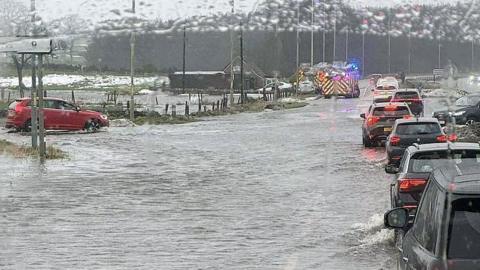 Traffic queues along a flooded road as cars drive through standing water, with emergency vehicles flashing blue lights in the distance during heavy rain.