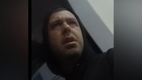 Image shows Watson being arrested. He has dark hair, and is looking up at the police officer's body-worn camera.