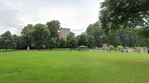 A Google Street View of a city park. Trees are along the edge of the park and a multi-storey building is visible beyond. A large grass area is in the foreground, and a playground with wooden structures is behind it.