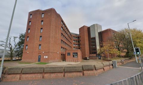 A picture of a multi-storey redbrick apartment block with traffic lights on the road outside the building.