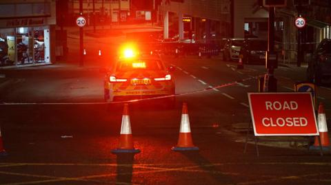 A police car is parked by a police cordon at night with its lights on. 