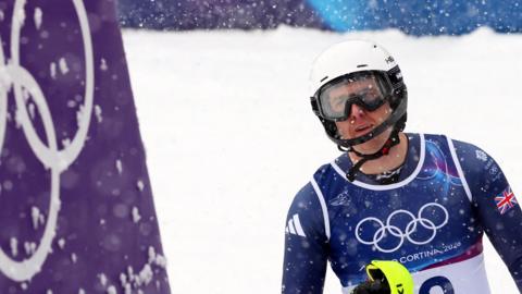 Team GB's Dave Ryding after his first run in the men's slalom in the Milano Cortina 2026 Olympics at Stelvio Ski Centre, Bormio, Italy. He is wearing Team GB's ski suit and white ski helmet. The Winter Olympics logo is to the side of him. 