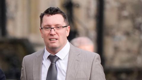 A man wearing a pale grey striped suit jacket, white shirt, metallic grey tie, and half-rimmed glasses walks along against a blurred outdoor background. His mouth is slightly open. He has brown hair which is receding. 