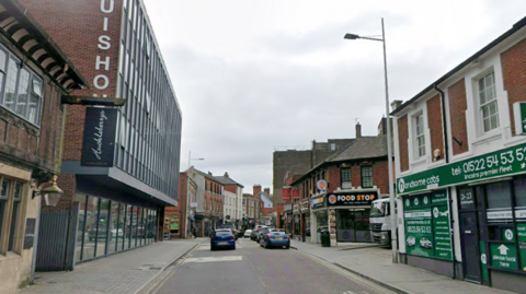 A view of Clasketgate in Lincoln. A large glass-fronted building is on the left, with a green taxi firm on the right. There is also a takeaway next to the taxi premises and cars are driving along the road.