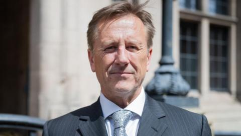Martin Murray is standing infront of the ocunty council buildings in Stafford looking at the camera, he is wearing a grey tie, shite shirt and black suit jacket. 